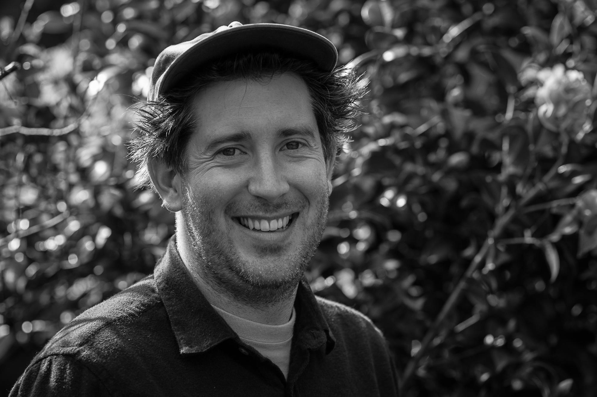 man with hat smiling and a leafy background (in black and white)
